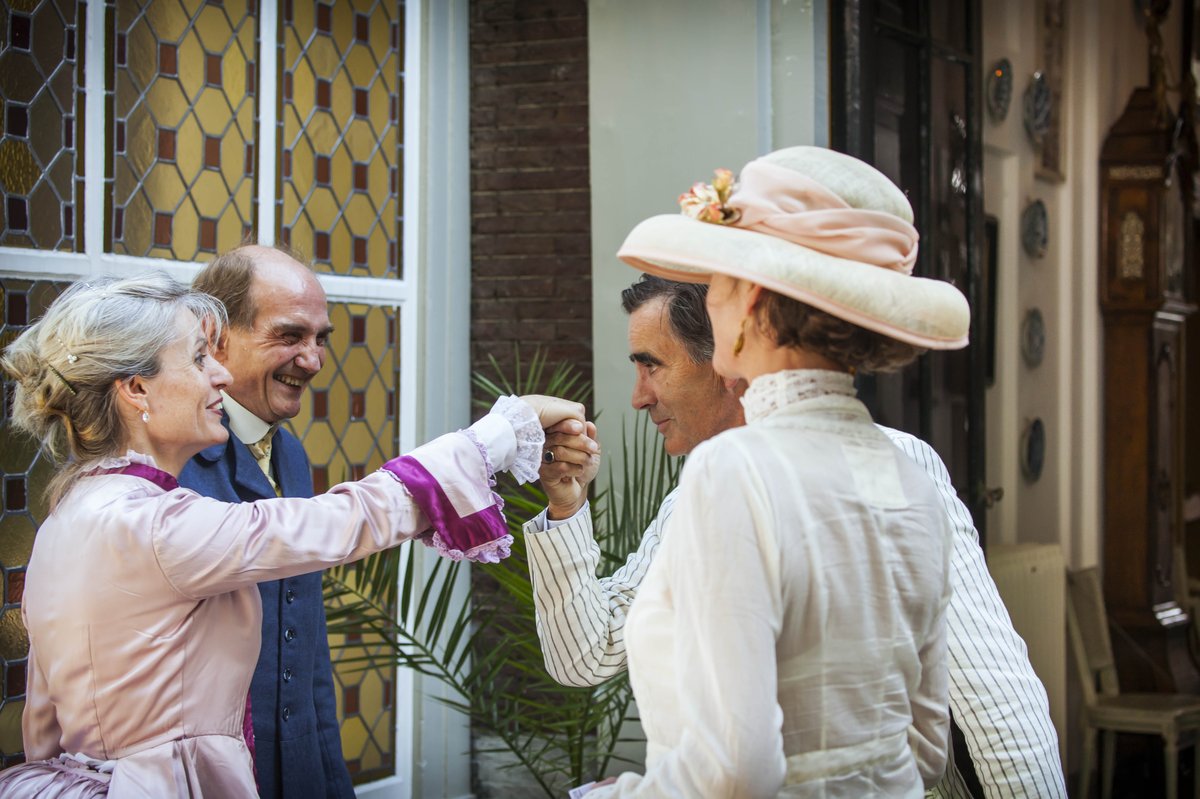 Een man staat op het punt mevrouw Van Gijn een handkus te geven. Hij kijkt meneer aan. Naast hem staat een vrouw in 19de eeuwse jurk met hoed.