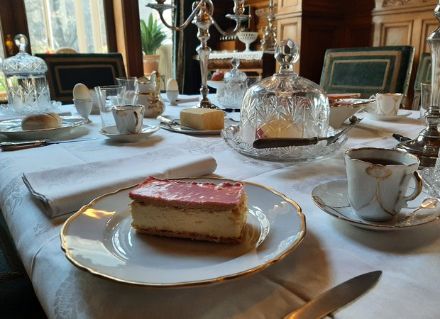 Op de rijk gedekte eettafel staat een wit servies met gouden randje. Op het bordje ligt een tompoes.