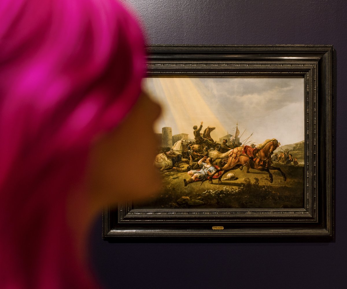 Vrouw met roze haar staat in een museumzaal. Achter haar een schilderij van Cuyp.