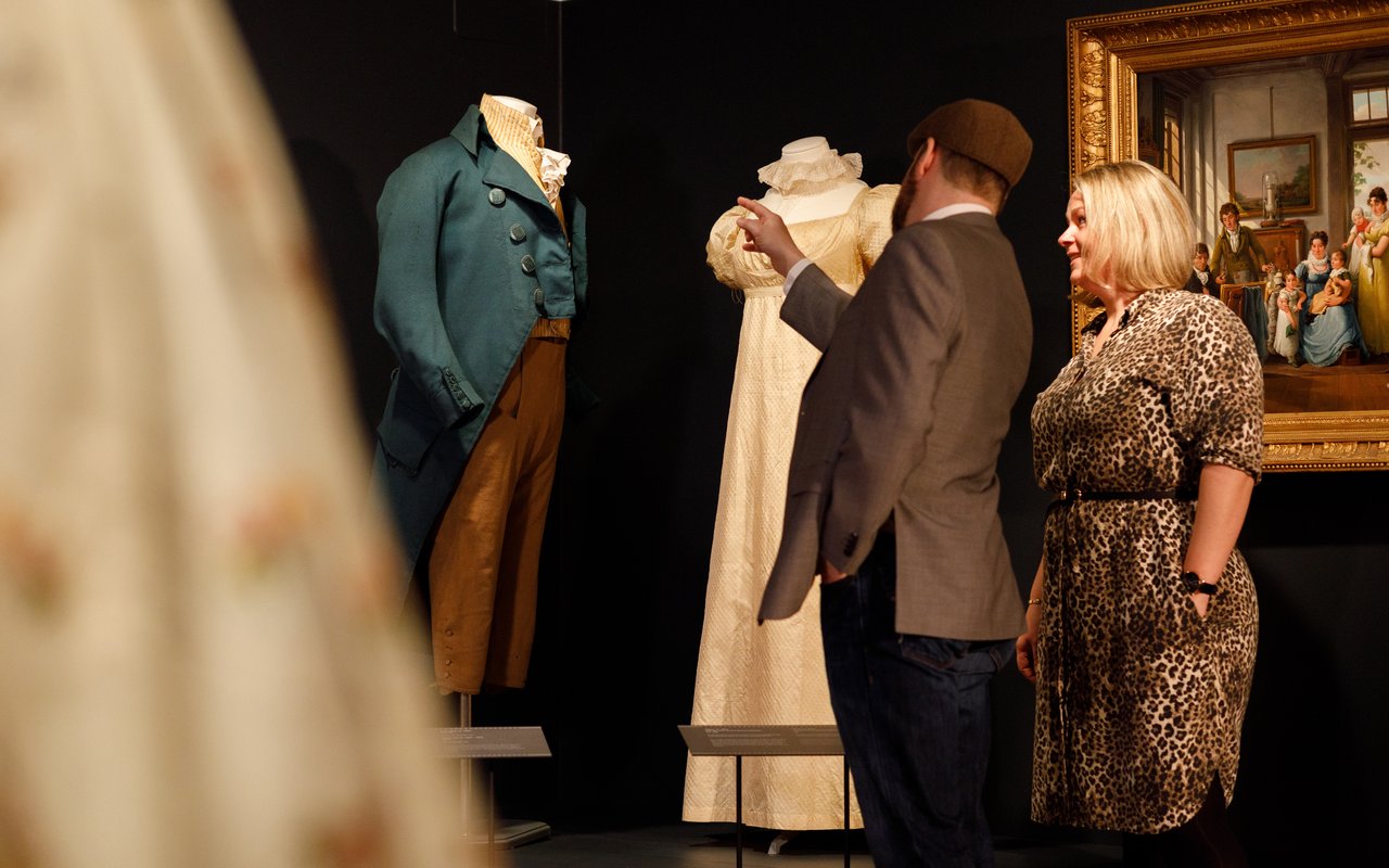 Een man en vrouw kijken naar een 19de eeuws pak en jurk die zijn opgesteld in het Dordrechts Museum.