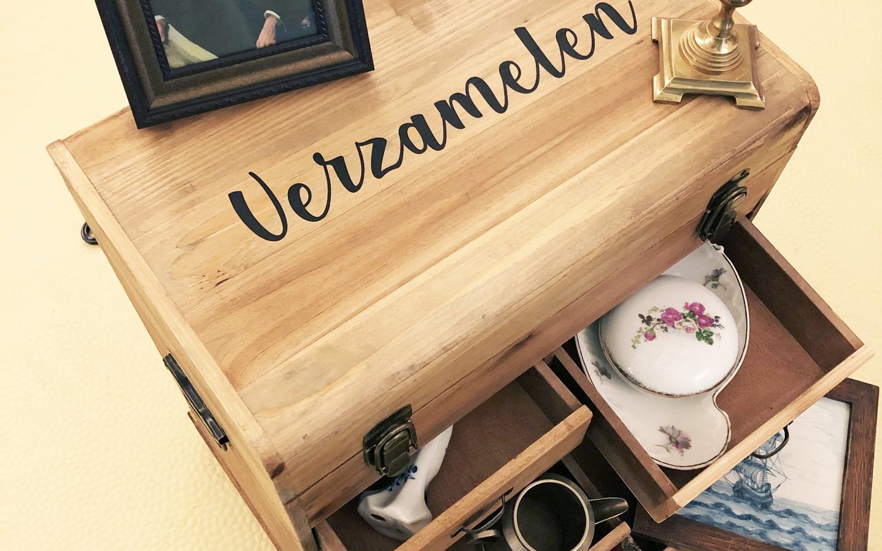 Een oude houten reiskoffer met open lades met daarin oud servies en andere items uit het Huis Van Gijn. Het portret van Simon staat boven op de koffer