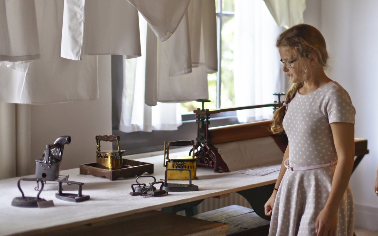 Een meisje staat in de waskamer van museum Huis Van Gijn. Ze kijkt naar oude strijkijzers op een tafel.