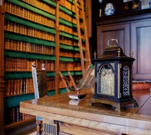 Bibliotheek. Er staat een trapje tegen de boekenwand. Op het houten bureau staat een klok.