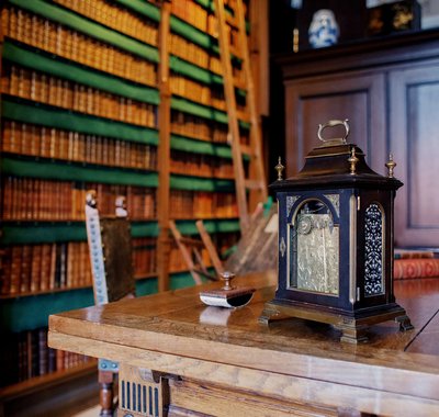 Bibliotheek. Er staat een trapje tegen de boekenwand. Op het houten bureau staat een klok.