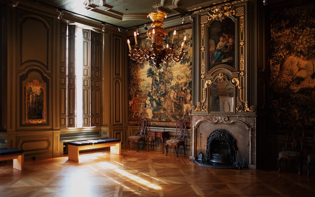 Zaal met houten vloer waarvan de muren zijn bedekt met wandtapijt. Er is een schouw en licht schijnt door het raam.