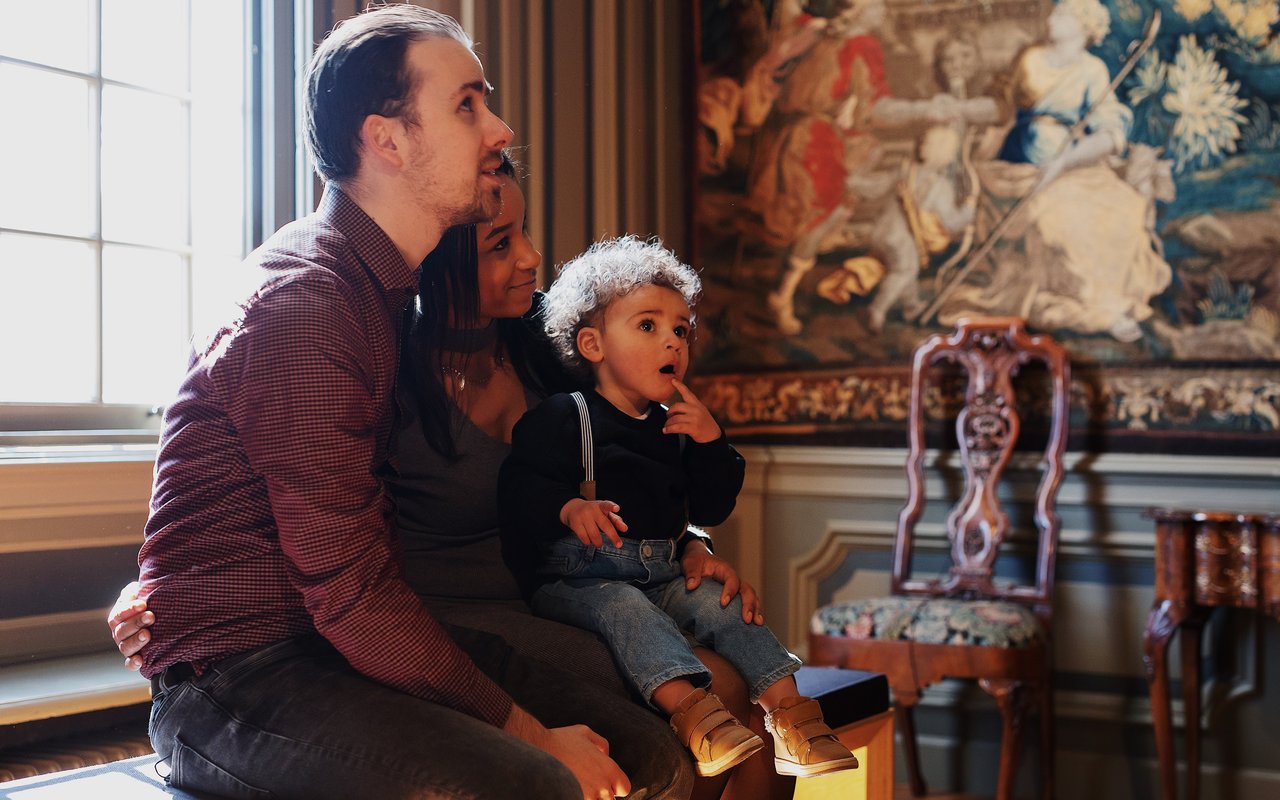 Een man en vrouw met peuter op schoot zitten op een bankje in de Tapijtenzaal. De peuter kijkt verbaast.