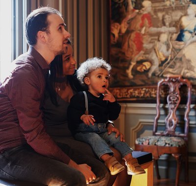 Een man en vrouw met peuter op schoot zitten op een bankje in de Tapijtenzaal. De peuter kijkt verbaast.