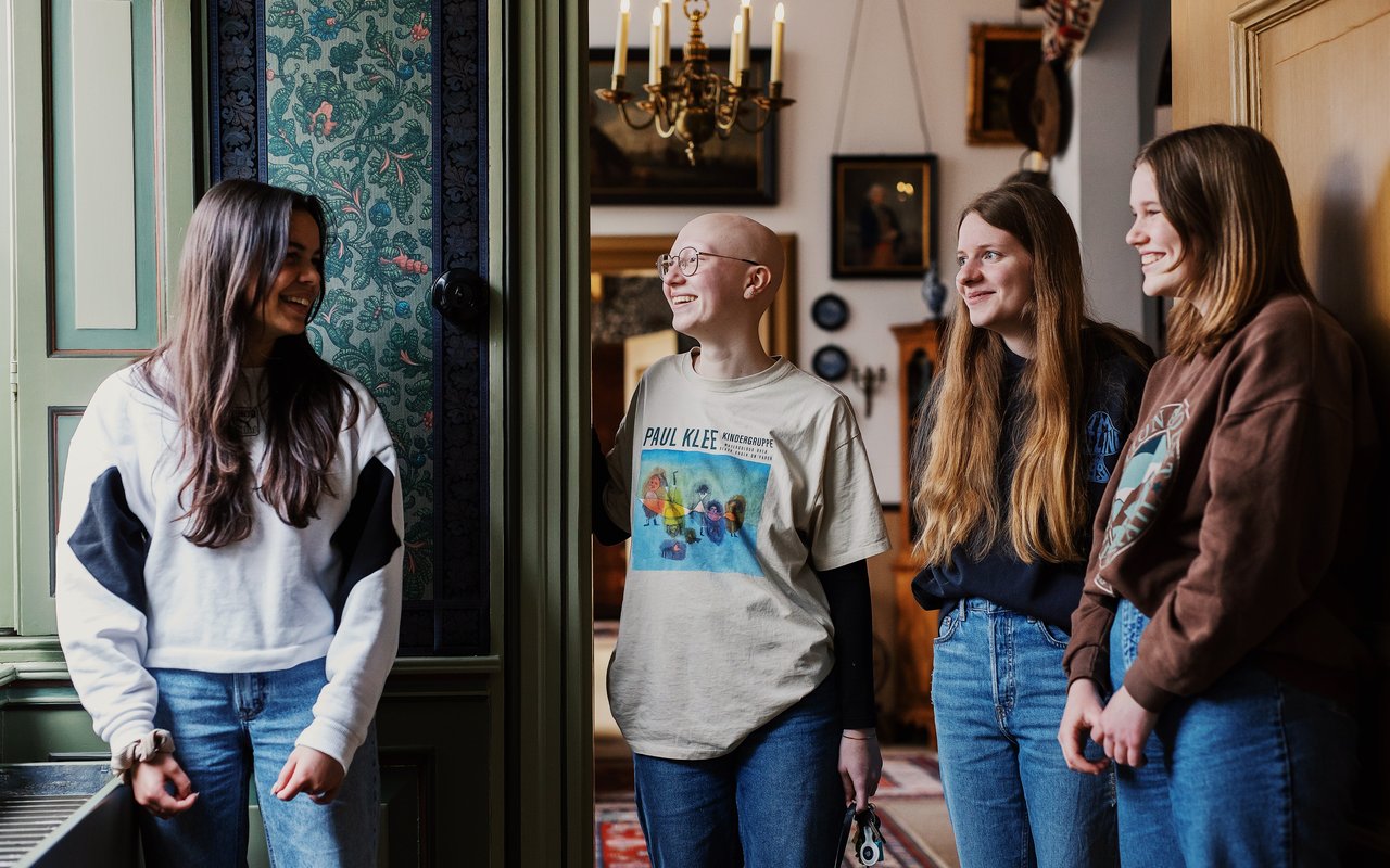 Vier jonge vrouwen staan in de gang van Huis Van Gijn. Ze lachen en praten met elkaar.