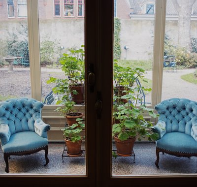 Doorkijkje naar de serre. Er staan twee met blauwe stof beklede stoelen, planten en een grote vaas.