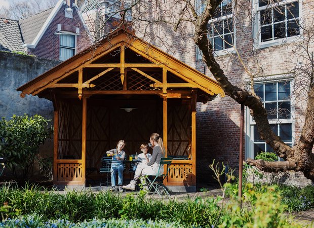 In de tuin zitten een vrouw met twee kindjes in het tuinhuisje te eten en te drinken.