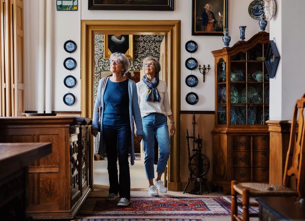 Twee vrouwen lopen door de gang op de eerste verdieping van Huis Van Gijn. Ze kijken om zich heen.