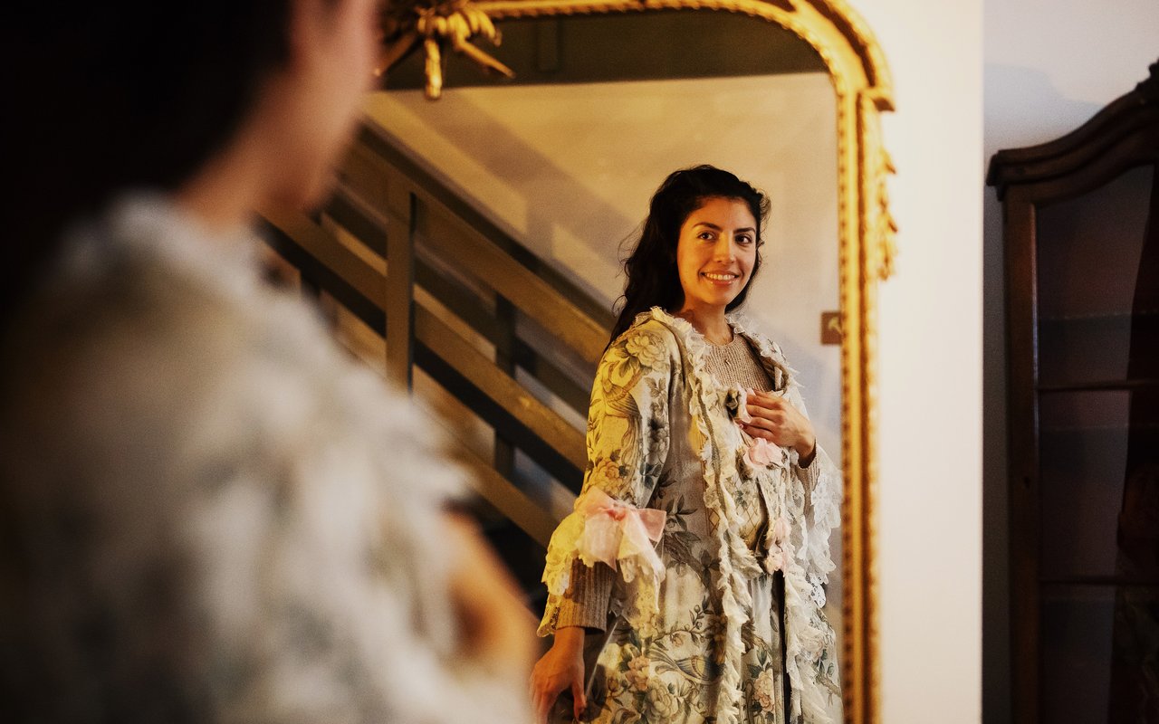 Verkleedzolder in Huis Van Gijn. Een vrouw kijkt vrolijk in de spiegel terwijl zij verkleed is in oude kledij uit 20e eeuw.