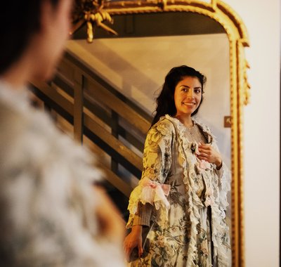 Verkleedzolder in Huis Van Gijn. Een vrouw kijkt vrolijk in de spiegel terwijl zij verkleed is in oude kledij uit 20e eeuw.