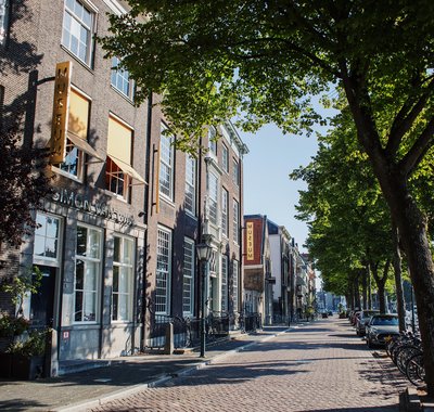 De straat en buitenkant van Huis Van Gijn. Het is lente, de zon schijnt en de bomen zijn groen. Er zijn geen mensen op straat.