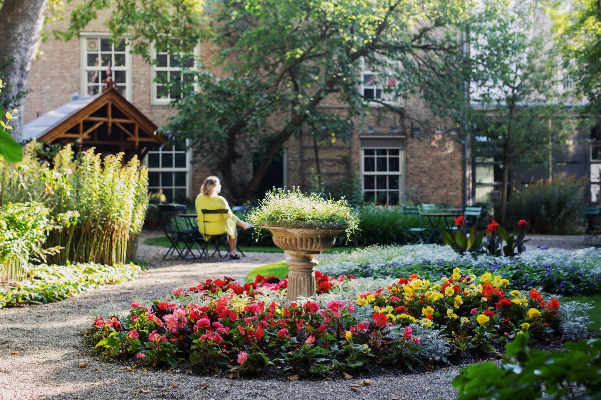 Zomertuin met bloemenperkje. Er zit een vrouw in gele jurk op een stoeltje in de zon.