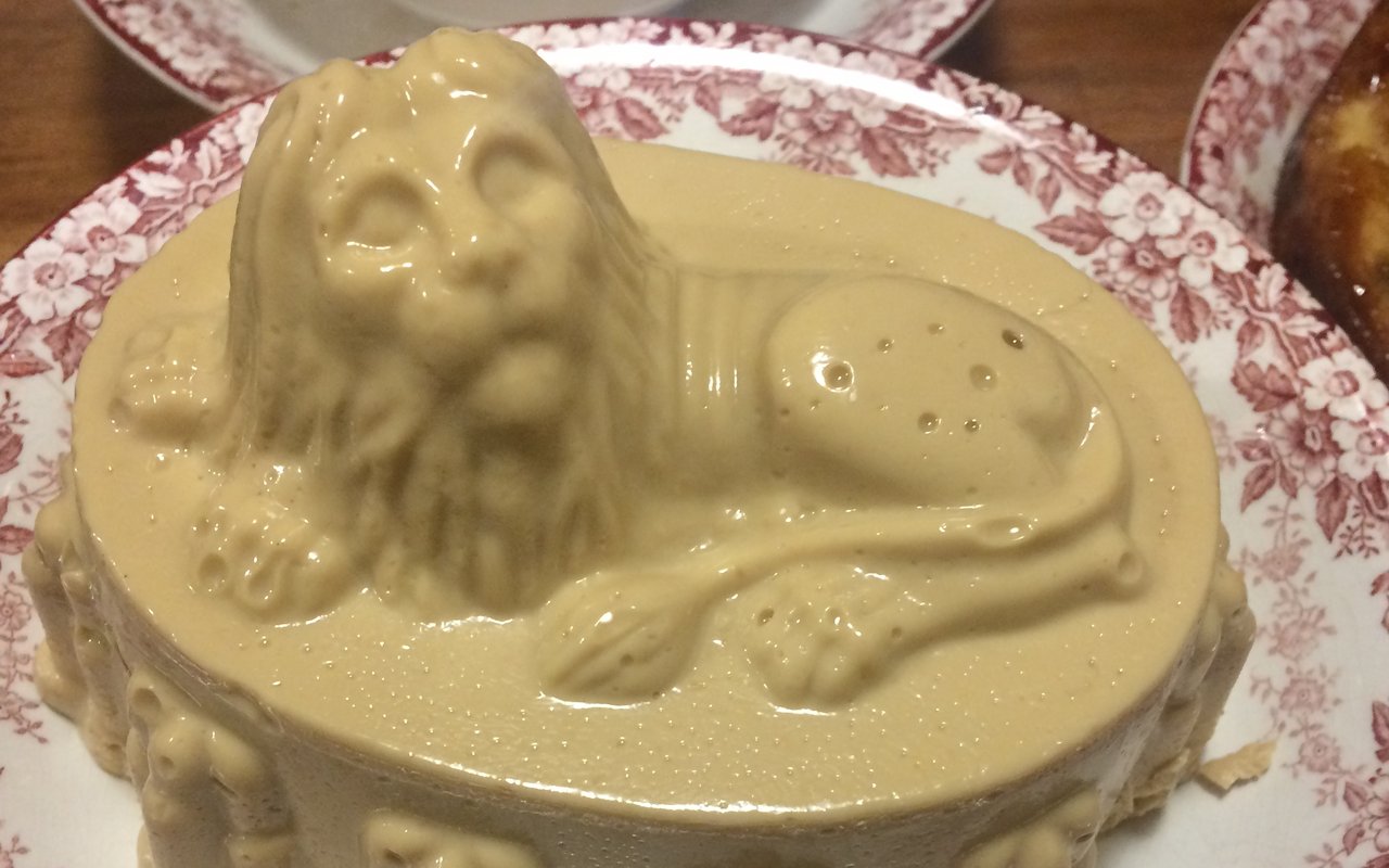 Een hopjespudding in de vorm van een liggende leeuw. De pudding ligt op een wit bord met rood bewerkte rand.