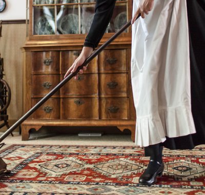 Een dienstmeisje gaat met een rolveger over een tapijt in Huis Van Gijn heen.