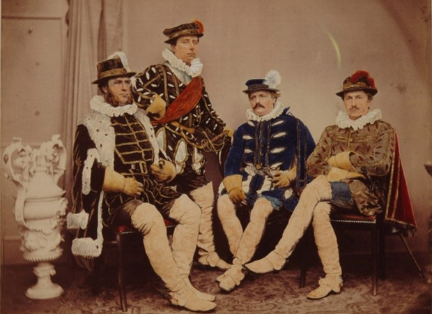 Ingekleurde foto van 4 zittende mannen in kostuum. Ze kijken ernstig. Naast hen staat een witte vaas.